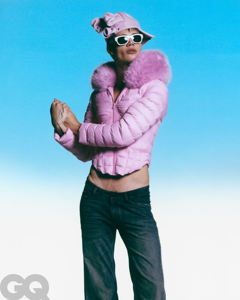 Person in pink puffer jacket, jeans, sunglasses, and hat poses against a light blue gradient backdrop in a fashion-style editorial shot.