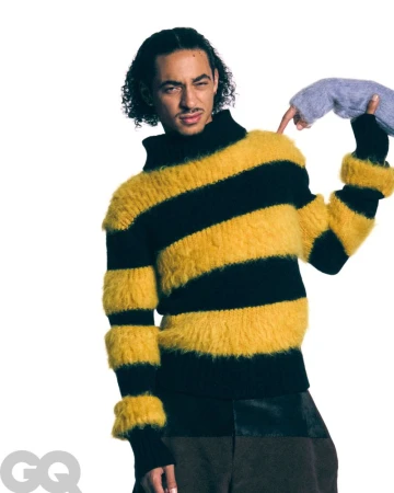 Man in a black-and-yellow striped furry sweater. Someone is touching his shoulder with a gray furry glove in a studio environment.