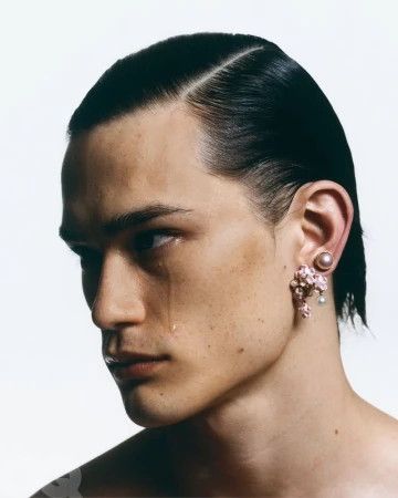 Close-up portrait of a young adult male with slicked-back black hair, a tear on his cheek, and statement floral earrings against a white backdrop.