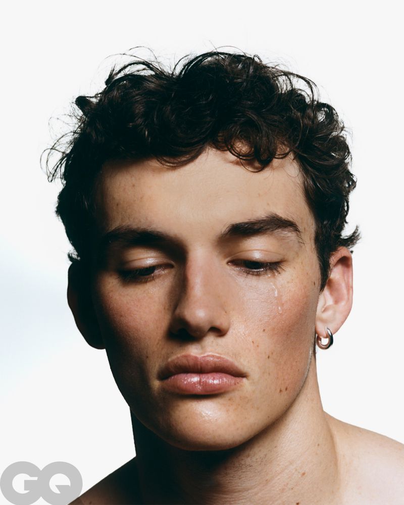 Close-up portrait of a young adult male with dark curly hair and a tear on his face, wearing a silver hoop earring, against a white backdrop.