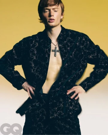 Red-haired adult male model posing in patterned jacket and pants with open chest, wearing necklace against a yellow backdrop.