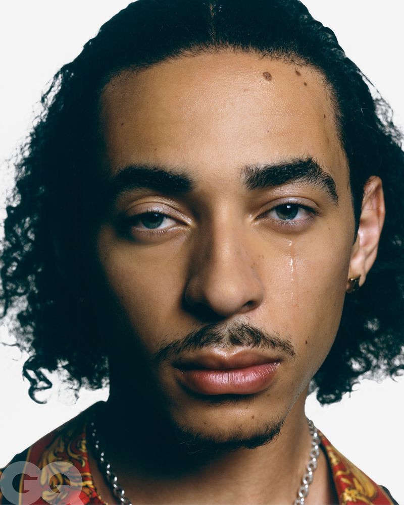 Close-up portrait of a young adult male with light skin and dark curly hair wearing a patterned shirt and chain necklace on a white background.