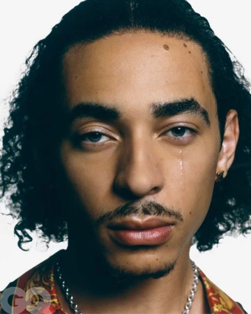 Close-up portrait of a young adult male with light skin and dark curly hair wearing a patterned shirt and chain necklace on a white background.