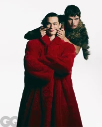 Two male models posing against a white backdrop, one wearing an oversized red plush coat while the other pinches his cheeks.