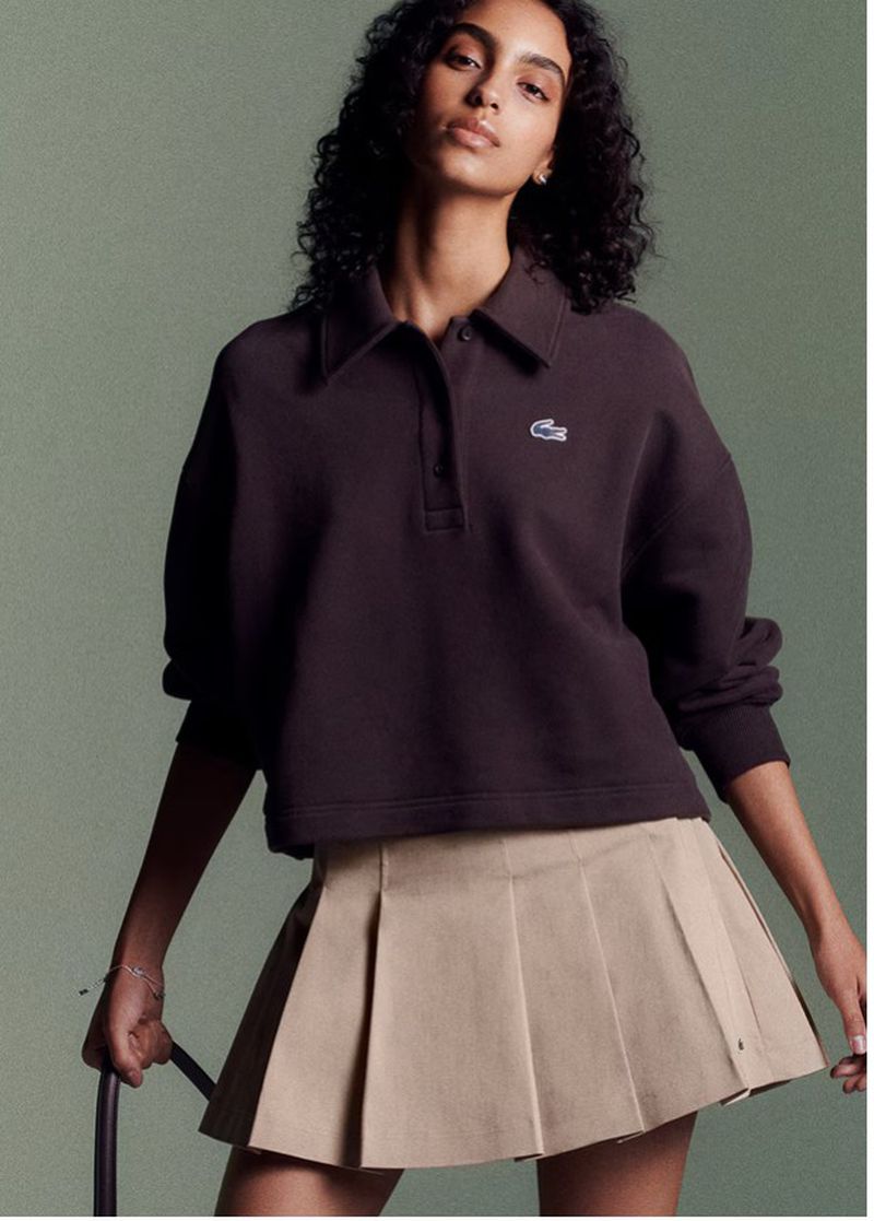 A brunette female fashion model poses indoors wearing a brown Lacoste logo sweatshirt and beige pleated miniskirt, studio shot.