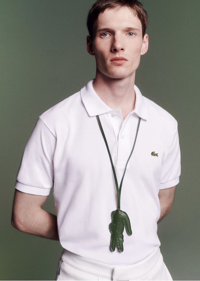 Male model in a white Lacoste polo shirt wearing a green crocodile leather lanyard pendant against a muted olive green studio backdrop.