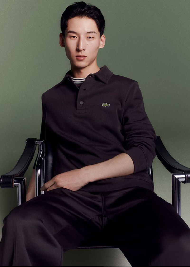 Portrait of an Asian male model in a dark polo shirt and pants, sitting on a chair in a studio with a green background.