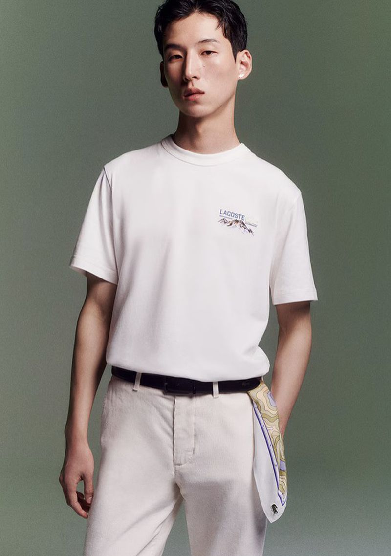 Model Woosang Kim in a white Lacoste t-shirt and cream trousers with a patterned scarf against an olive green backdrop.