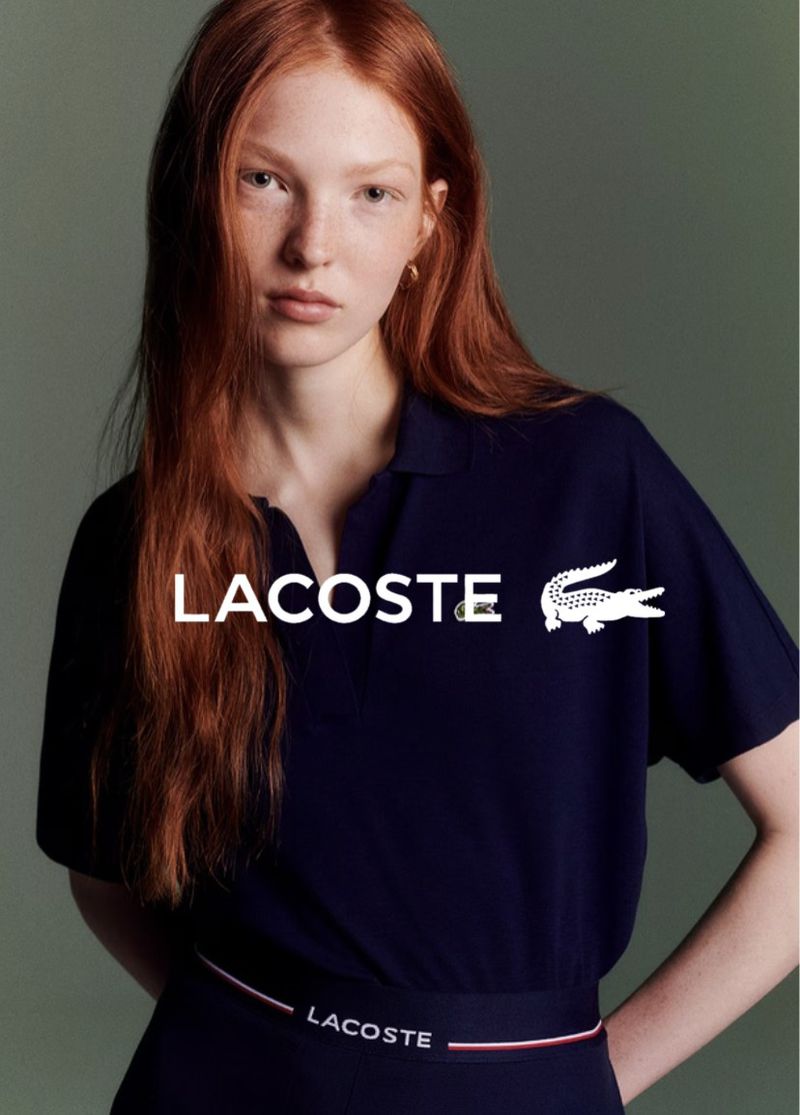 A young woman with long red hair is featured in a fashion shot, wearing a Lacoste polo dress against a muted grey background.