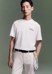 Model Woosang Kim in a white Lacoste t-shirt and cream trousers with a patterned scarf against an olive green backdrop.
