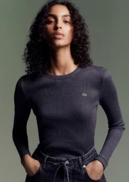 Model with curly dark hair wearing a charcoal grey long-sleeve ribbed top and black denim against an olive green studio backdrop.