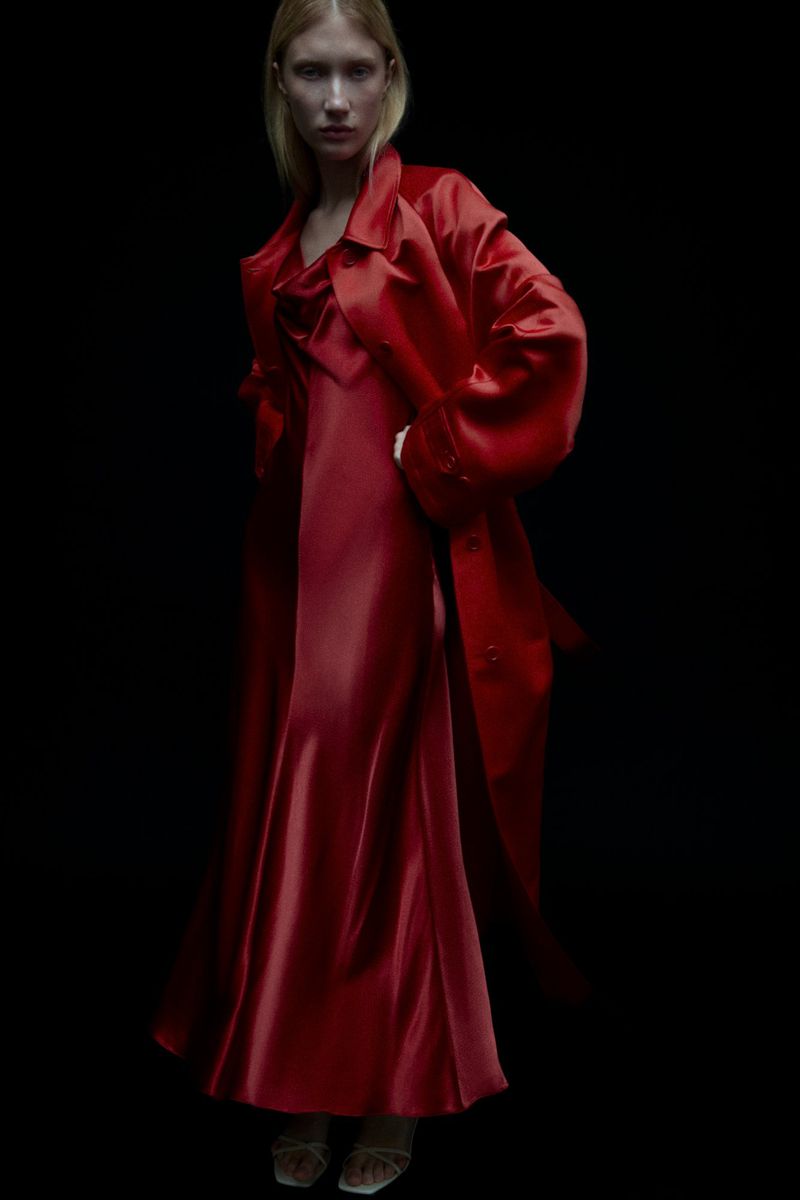 Fashion shot: A woman with fair skin and blonde hair is posing in a shiny red dress and a matching red overcoat against a dark backdrop.