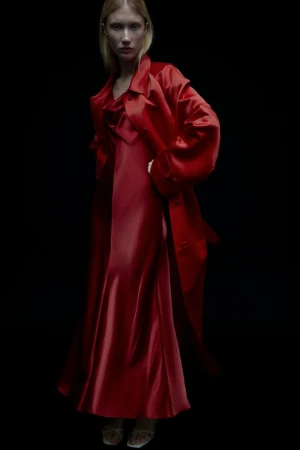 Fashion shot: A woman with fair skin and blonde hair is posing in a shiny red dress and a matching red overcoat against a dark backdrop.