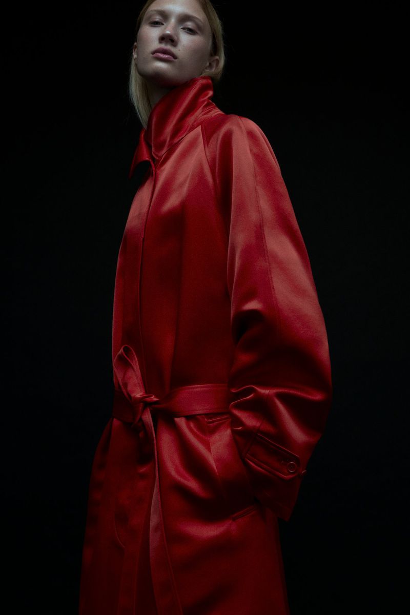 Adult female in a red satin trench coat against a dark background, medium shot, fashion editorial, dramatic lighting.