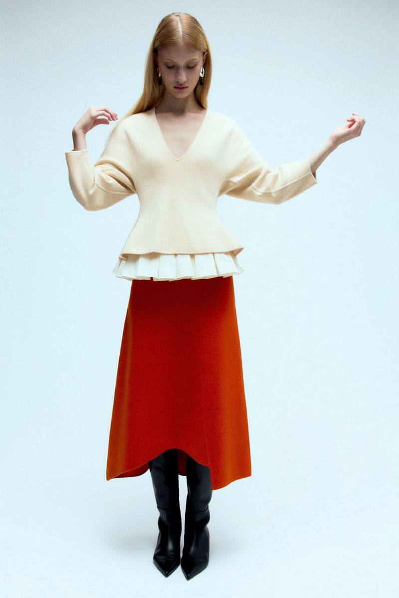 A fair-skinned woman poses in a studio wearing a cream sweater with a peplum waist, red midi skirt, and black pointed boots on a white background.
