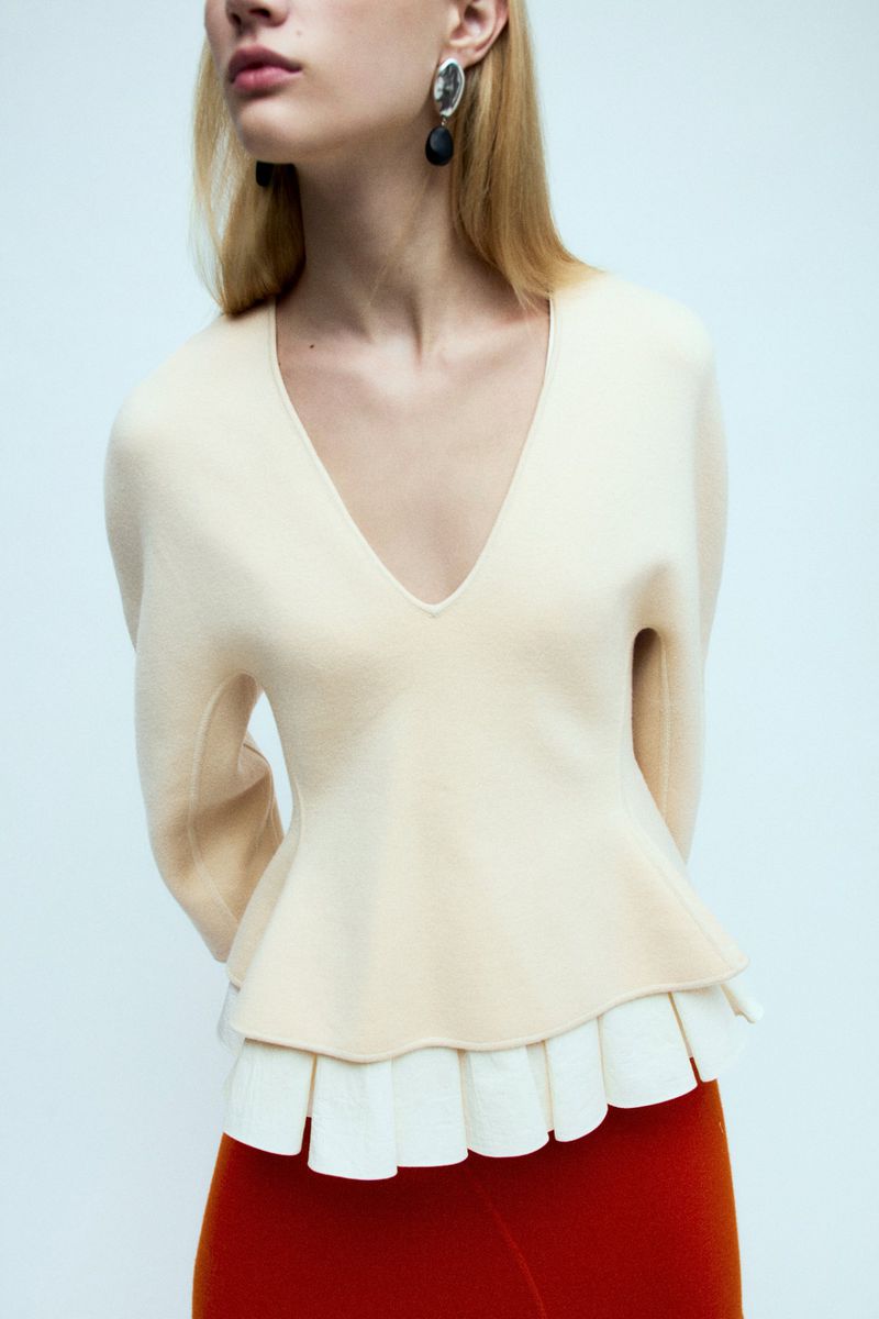 Woman wearing a V-neck sweater with pleated hem and red pants, posing indoors in a studio with a light background.