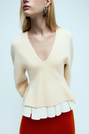 Woman wearing a V-neck sweater with pleated hem and red pants, posing indoors in a studio with a light background.