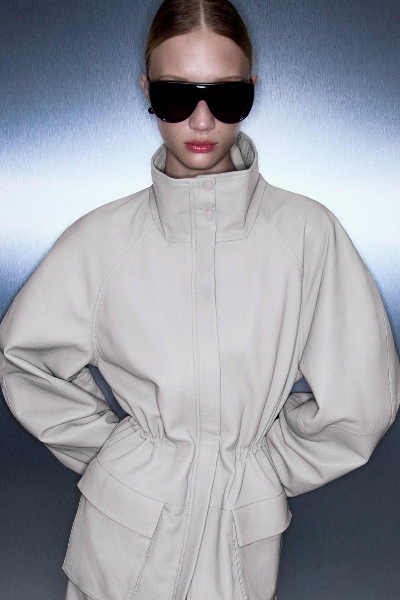 Fashion portrait of a woman wearing an off-white coat and black sunglasses, with a silver background. Studio shot, eye-level angle.