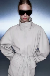 Model Daria Danilenko wearing an off-white cinched coat and black shield sunglasses against a cool metallic gradient backdrop.