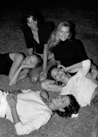 Five women pose on grass in a black and white photograph. The women are wearing casual tops, lying on the grass and smiling at the camera.