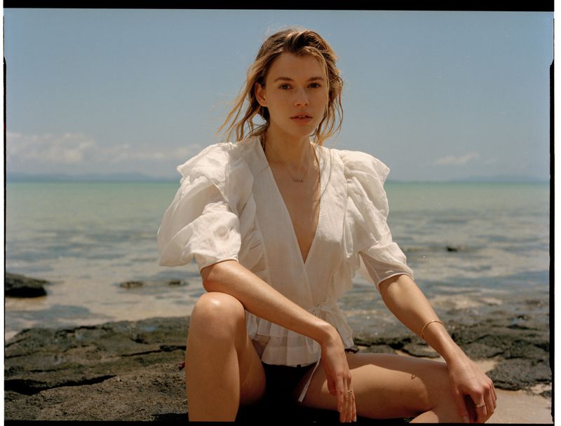 Woman in white blouse kneeling on dark rocky coast by ocean for Harper's Bazaar Australia editorial.
