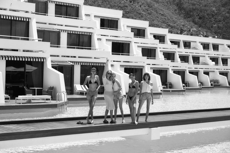 Five women pose together on a dock near white modern architecture and clear water, with a hillside in the background. Black and white.