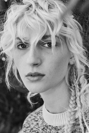 Close-up portrait of a woman with blonde curly hair and braids wearing a textured sweater