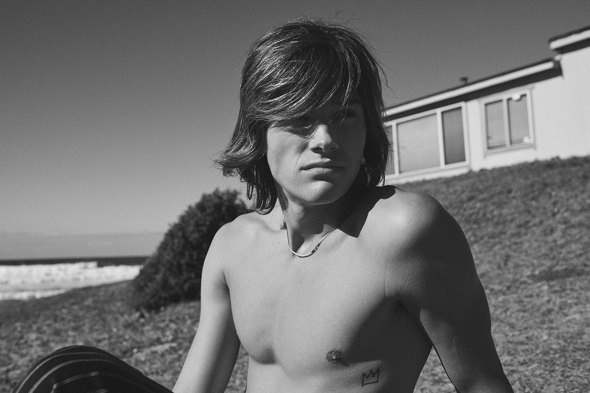 Black and white portrait of a shirtless young man with long hair and a tattoo sitting outdoors, with the beach and a house in the background.