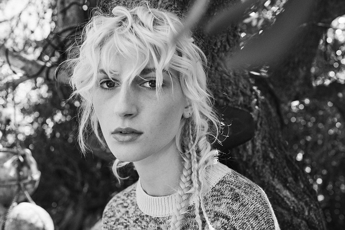 Black and white portrait of a blonde woman outdoors wearing a patterned sweater in front of tree foliage.