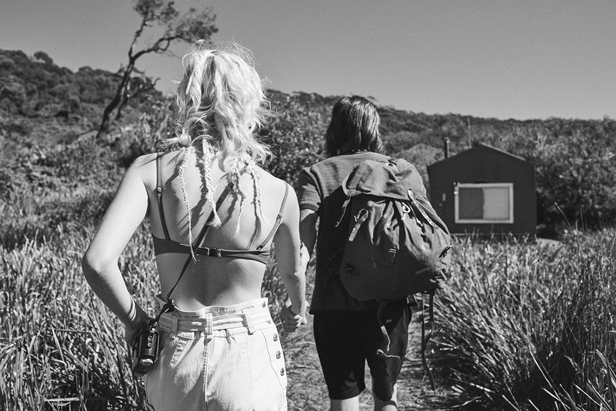 Two people hiking in a nature preserve, wearing casual clothing and carrying backpacks, with a cabin visible in the background on a sunny day.