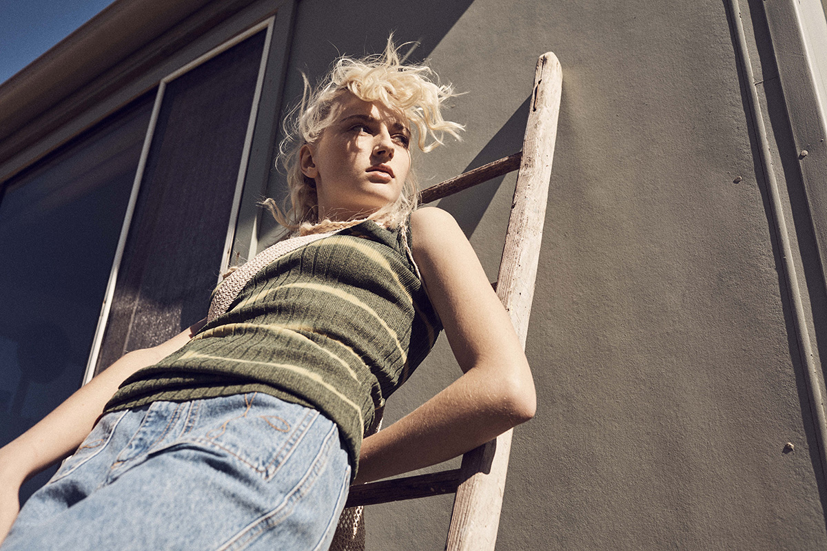 Model with blonde hair wearing a green striped top leaning against a wooden ladder on a building exterior