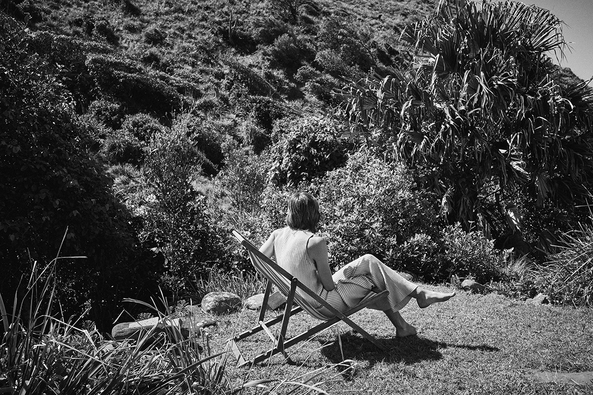 Adult woman relaxing in a wooden deck chair on grassy lawn in front of lush green hillside, sunny monochrome lifestyle image.