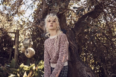 Blonde woman wearing a textured knit sweater leaning against an olive tree in an outdoor setting for Kaltblut