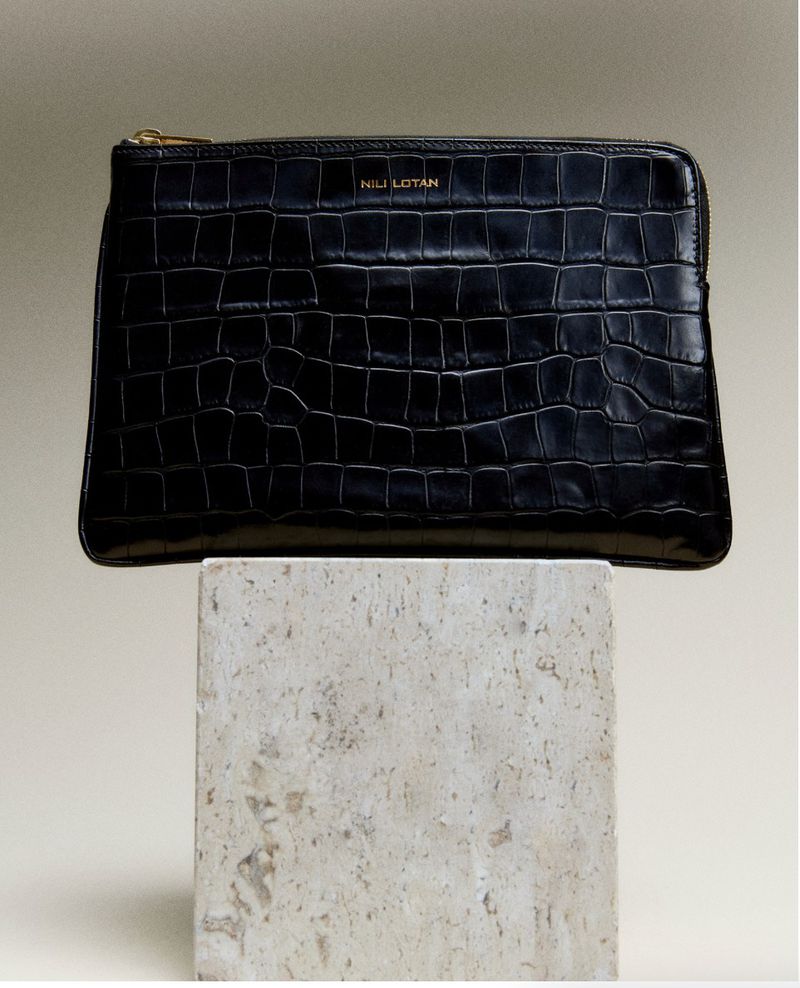 Stylish black leather clutch with a crocodile pattern, showcasing the Nili Lotan logo, resting on a textured stone block against a neutral backdrop.