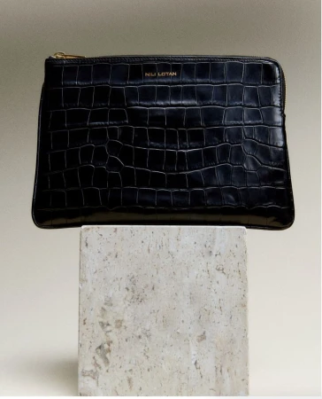 Stylish black leather clutch with a crocodile pattern, showcasing the Nili Lotan logo, resting on a textured stone block against a neutral backdrop.
