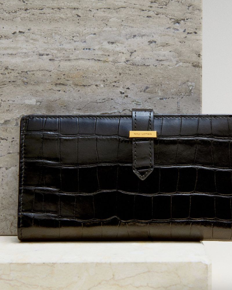 Close-up of a black wallet with a faux crocodile texture, featuring a gold-tone clasp, set against a marble surface and a travertine tile background.