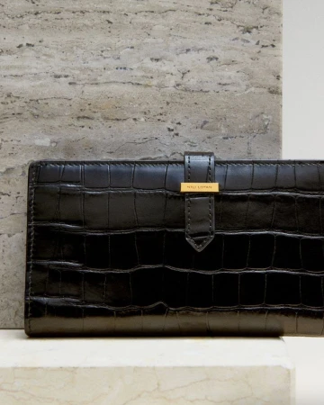 Close-up of a black wallet with a faux crocodile texture, featuring a gold-tone clasp, set against a marble surface and a travertine tile background.