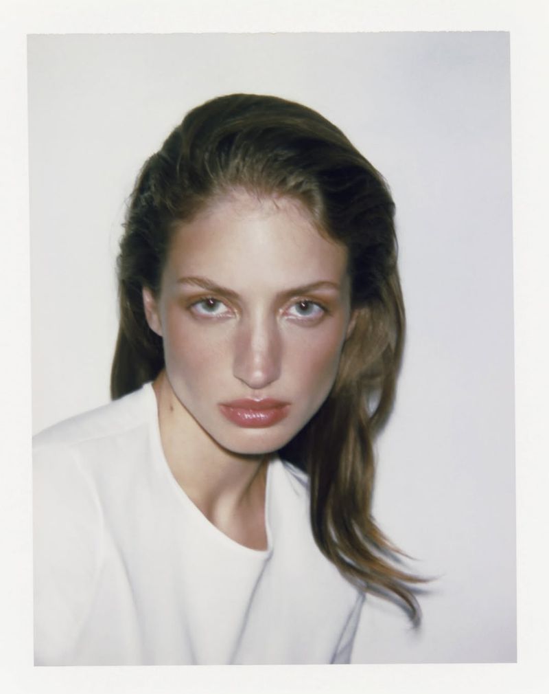 Close-up portrait of a young adult woman with long brown hair wearing a white top, set against a plain white background.