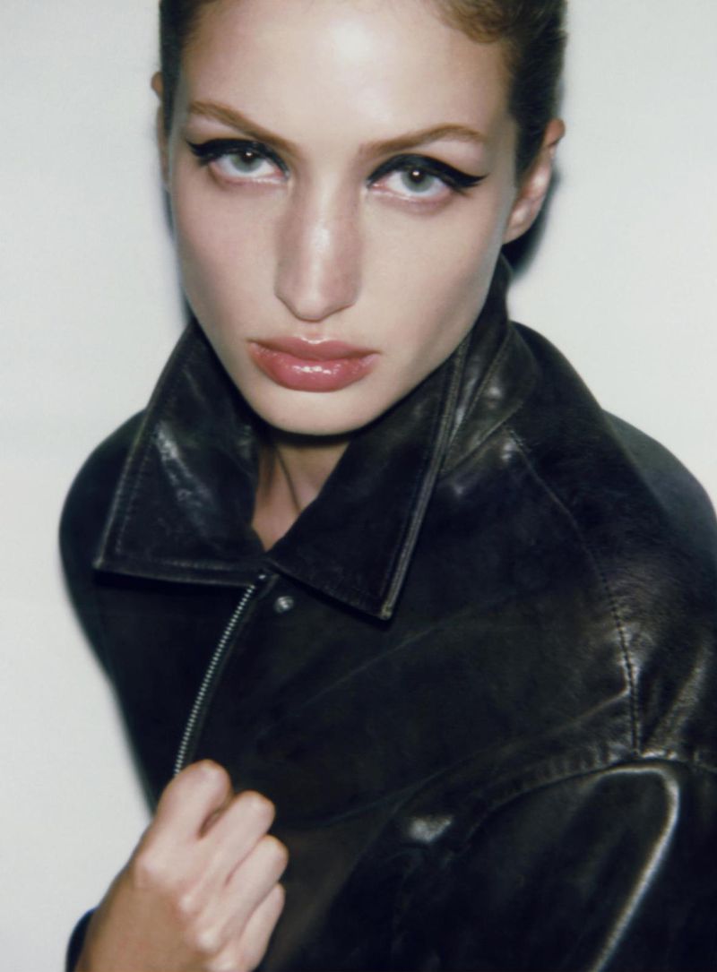 Close-up portrait of a woman with striking eye makeup wearing a black leather jacket against a neutral background, fashion shot.