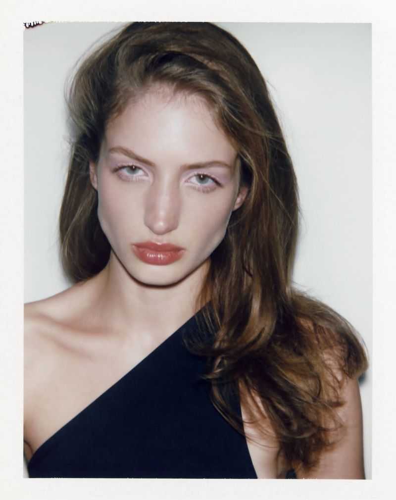 Portrait of a young woman with long brown hair wearing a one-shoulder black top against a white background in a close-up studio shot.