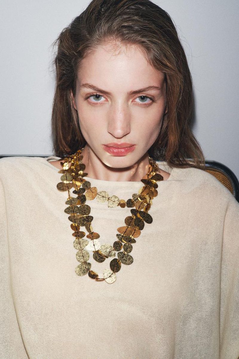 A young woman with brown hair and blue eyes is posing indoors, wearing a gold layered necklace and a light tan sweater.