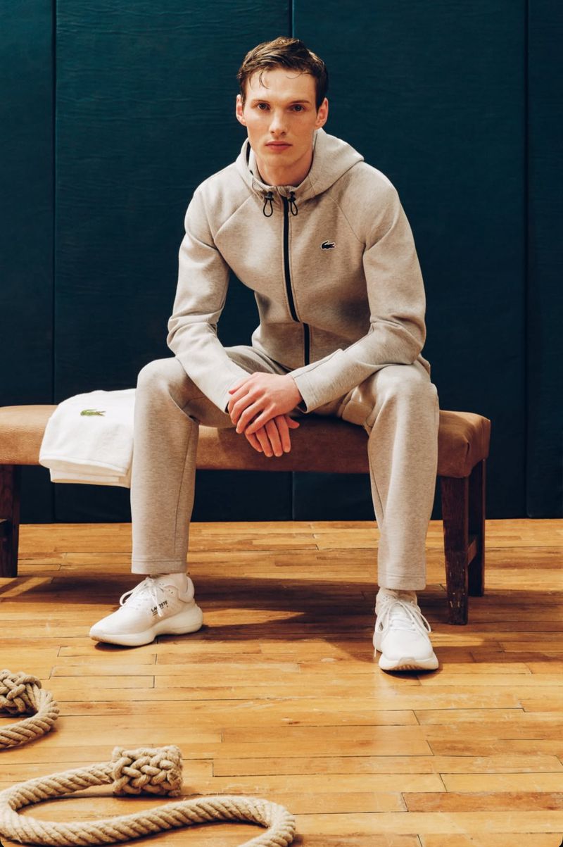 A young adult male in a grey hooded tracksuit sits on a bench in an indoor gym, with a rope on the floor and a dark green wall.