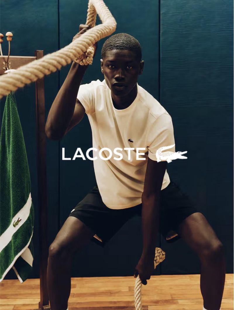 Athletic Black man works out with rope in a bright studio. He wears a white Lacoste shirt and black shorts. Towel and coat rack on the side.