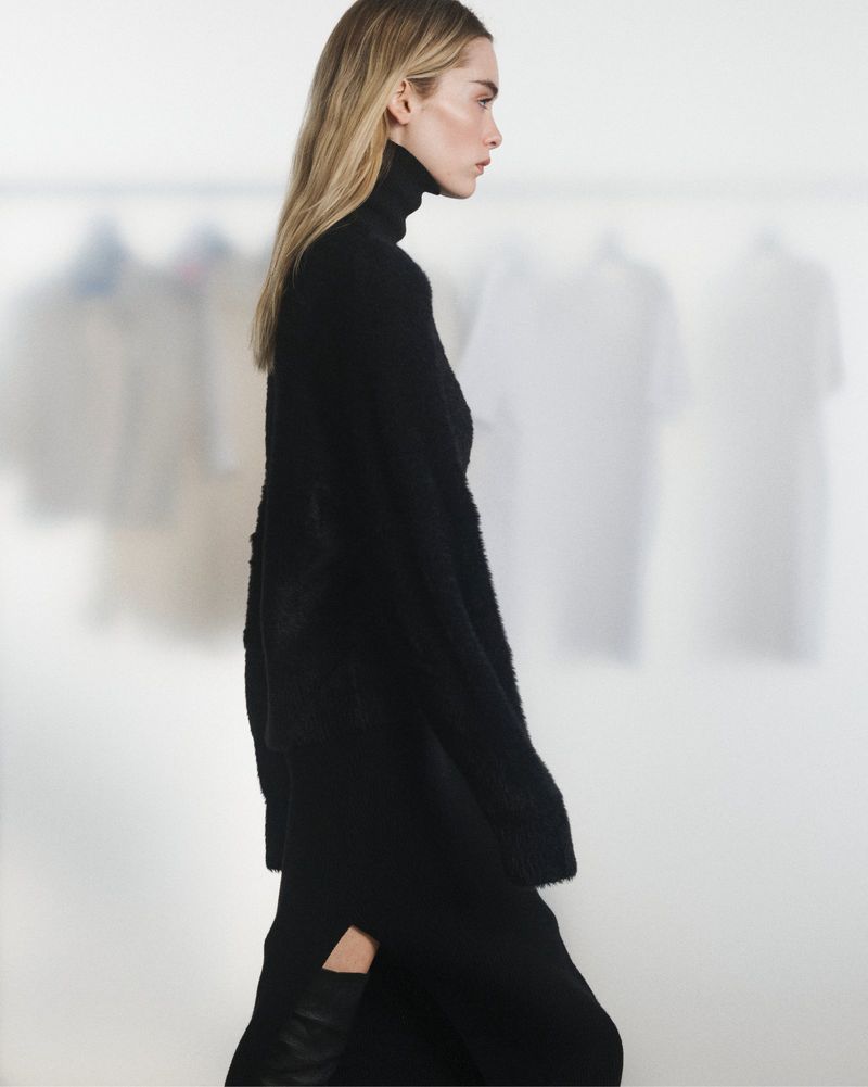 A side view of a blond adult woman in a black turtleneck sweater dress on a bright background with blurred garments. Fashion shoot.