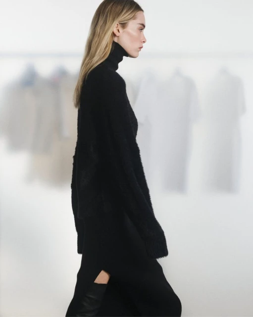 A side view of a blond adult woman in a black turtleneck sweater dress on a bright background with blurred garments. Fashion shoot.