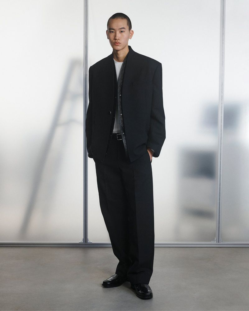 Model wearing an oversized black suit jacket and wide-leg trousers standing before frosted glass studio panels.