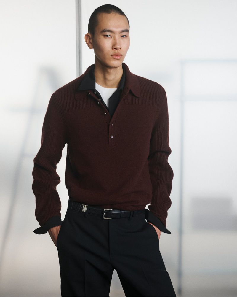 An Asian male model poses indoors wearing a maroon collared ribbed sweater, black trousers, and a black belt, against a blurred background.