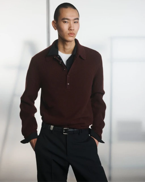 An Asian male model poses indoors wearing a maroon collared ribbed sweater, black trousers, and a black belt, against a blurred background.