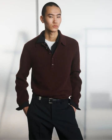 An Asian male model poses indoors wearing a maroon collared ribbed sweater, black trousers, and a black belt, against a blurred background.