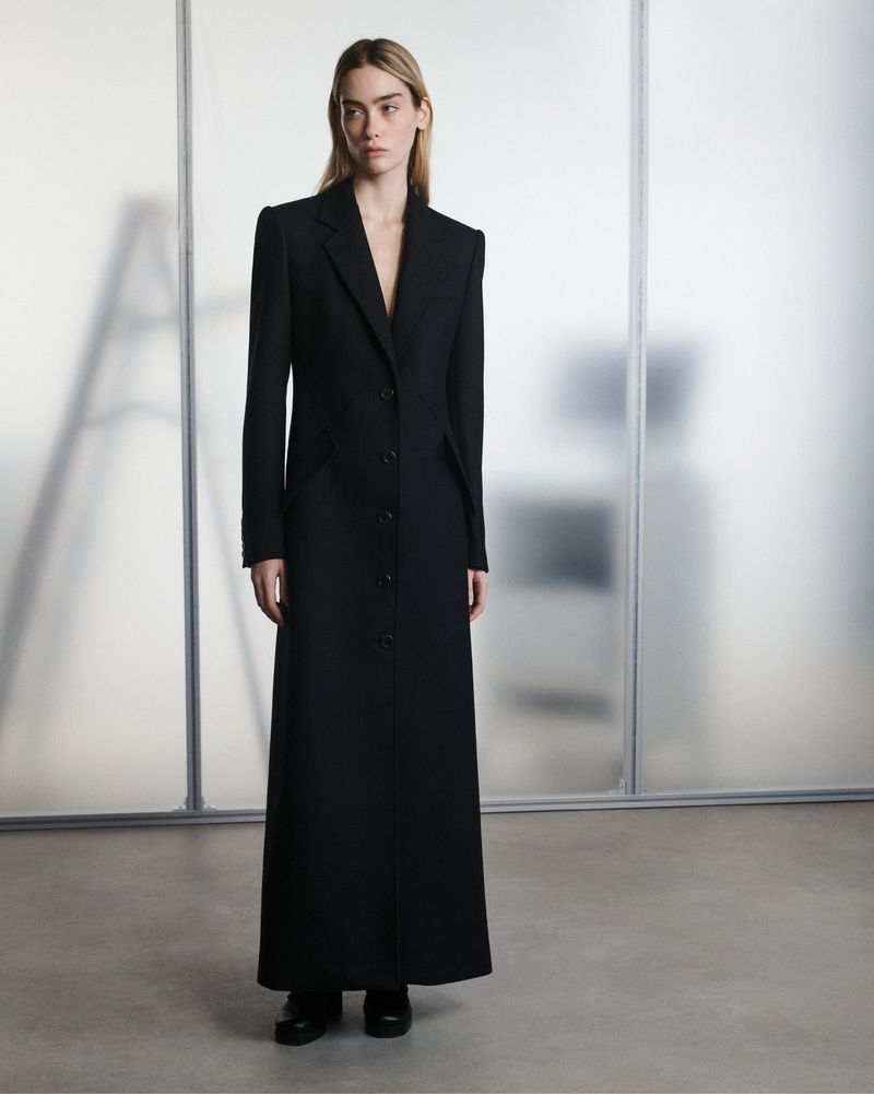 A woman in a long black coat poses indoors against a frosted glass wall, creating a minimalist and fashion-forward presentation.